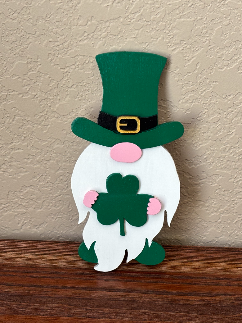 Decorative paper gnome with a green hat and pink cheeks holding shamrocks on a wooden surface.