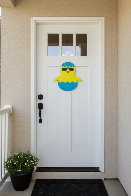 Decorative door hanger of a yellow chick in an egg with sunglasses and a blue cap on a brown door.