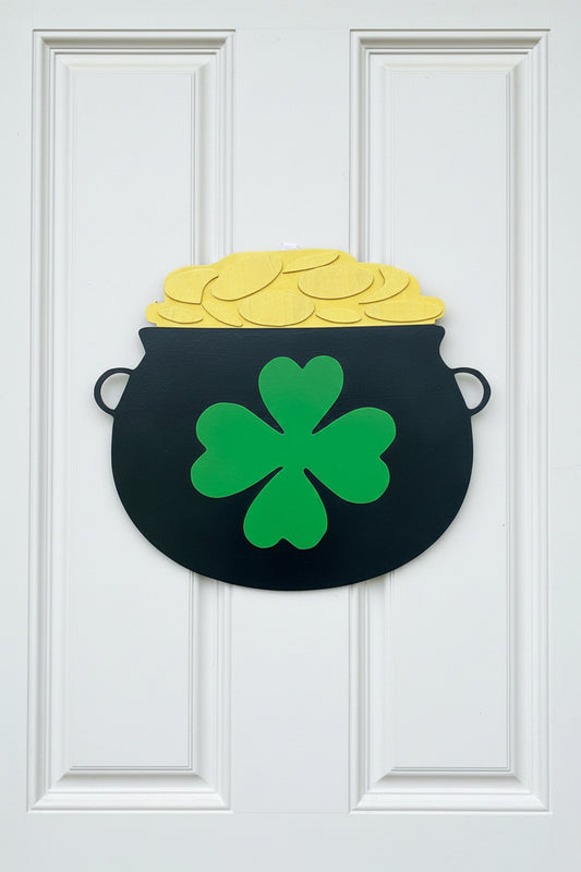 Decorative door hanger shaped like a pot of gold with a green four-leaf clover on a beige door.