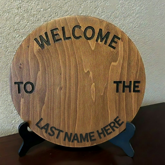 Hand-painted 12-inch customizable birch round with Welcome to the Last Name Here text, displayed on a black easel stand, handcrafted wood home decor